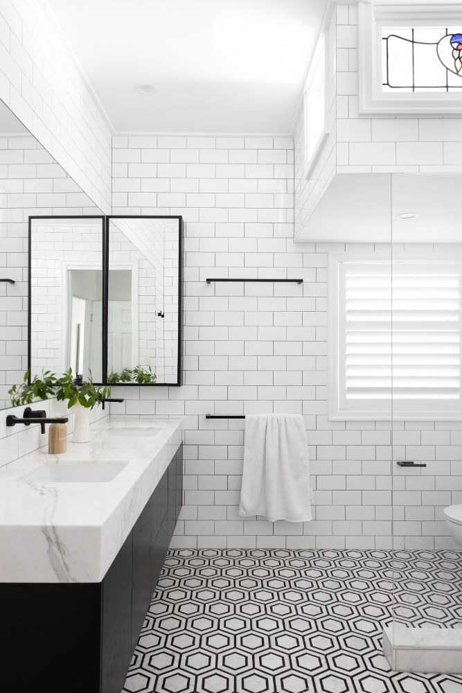A monochromatic modern bathroom showcasing traditional stained glass high windows. Black vanity, marble benchtop, black framed mirror and black and white hexagon marble floor.