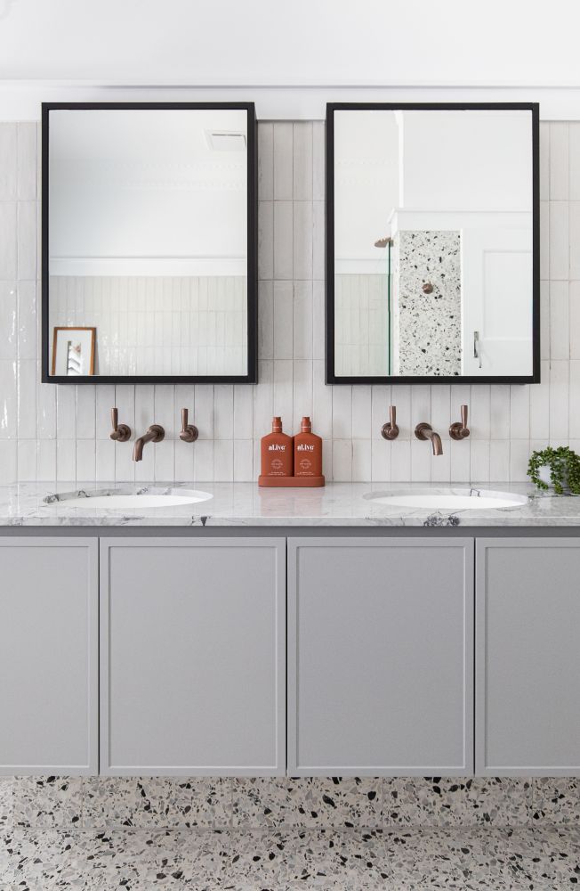 Sleek bathroom design featuring two black-framed mirrors, bronze fixtures, terrazzo flooring, and grey cabinetry complemented by white subway tile.