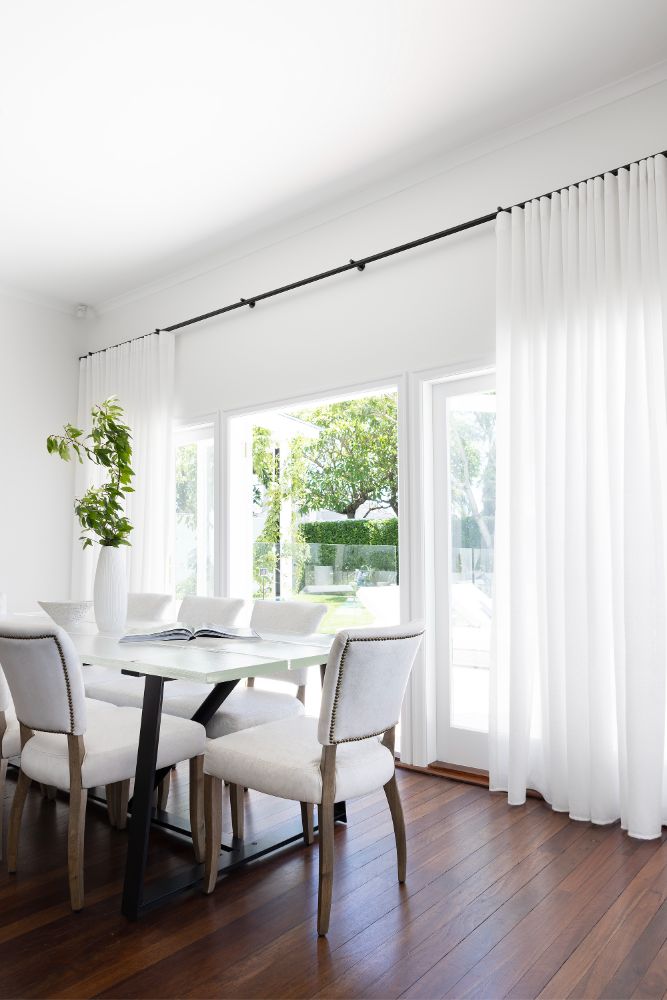 Elegant dining room with white sheer curtains, a modern black dining table, upholstered chairs, an indoor plant in a tall vase, and large windows revealing a lush garden view.