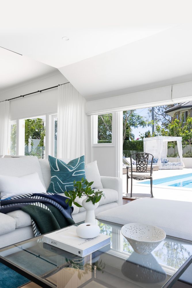 Modern coastal-themed living room with white sofas, teal-patterned pillows, a glass coffee table with decor and books, large windows showing an inviting pool area with an outdoor canopy lounge.