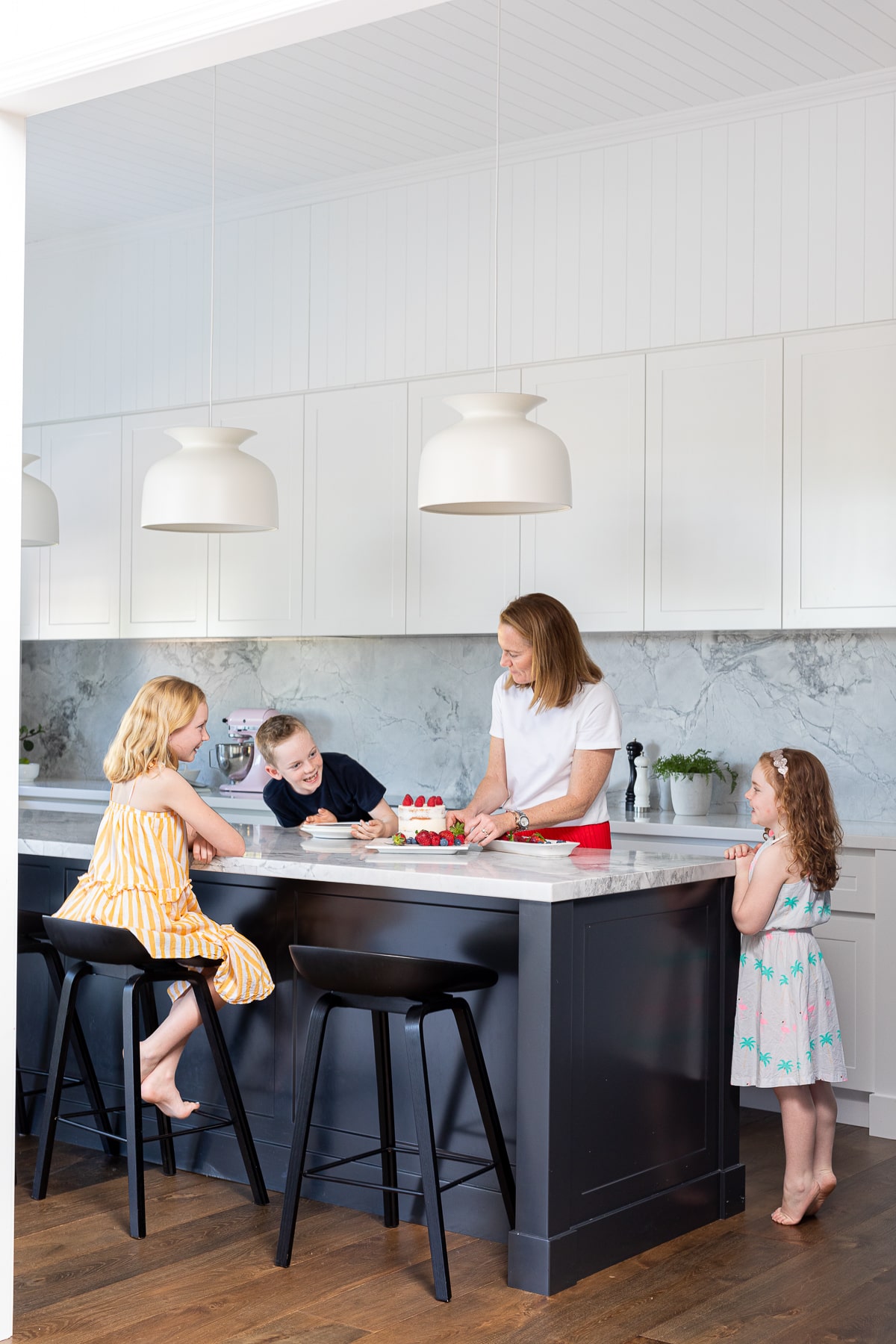 A woman and three young children gather around a black kitchen island as she serves cake and strawberries