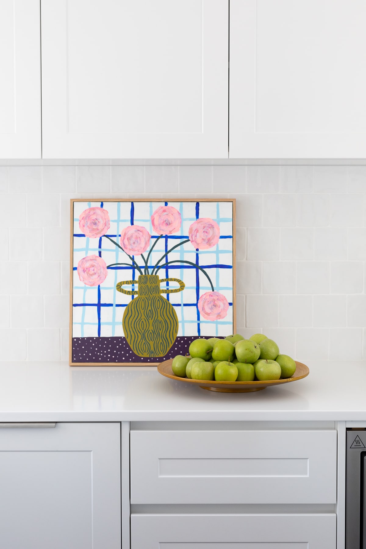 Contemporary kitchen decor featuring a colourful floral painting above a bowl of fresh green apples on a white countertop.