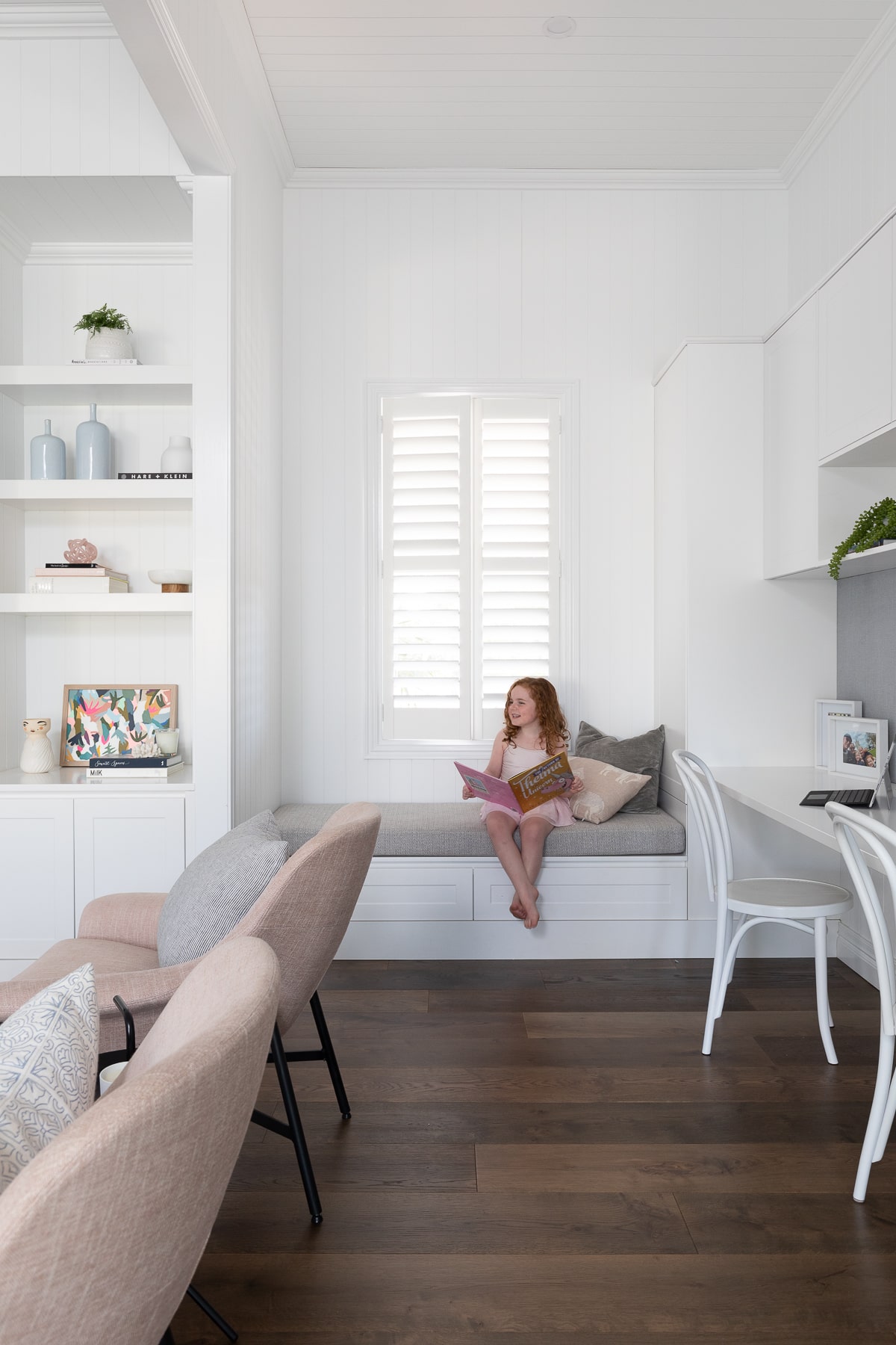 A young girl enjoys reading in a sunlit corner with built-in seating and open shelving, featuring elegant white decor and rich wooden flooring.