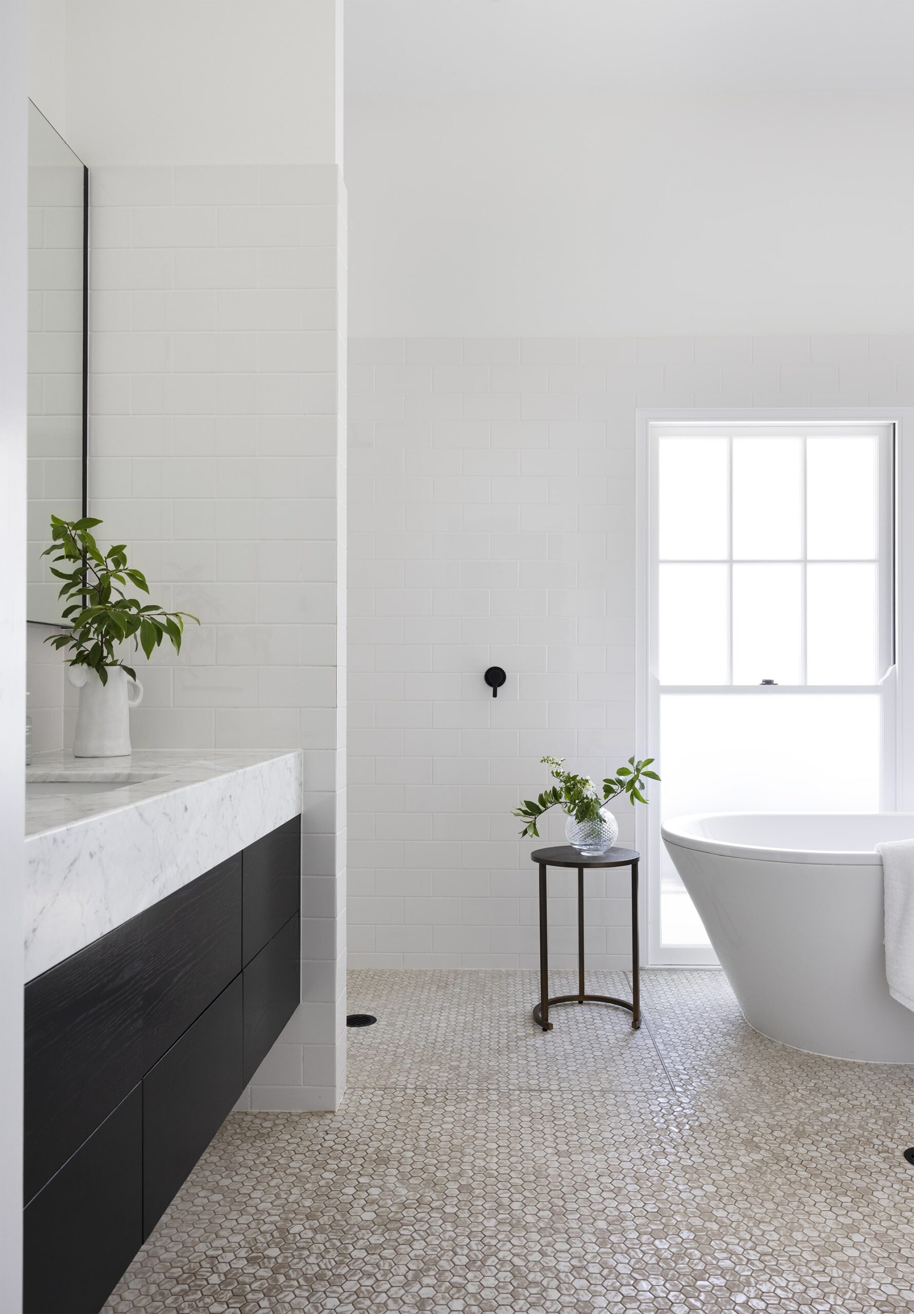 Modern bathroom with white subway tiles, marble countertop vanity, dark cabinetry, marble mosaic floor, and a free-standing bathtub by a window, accented with green plants.