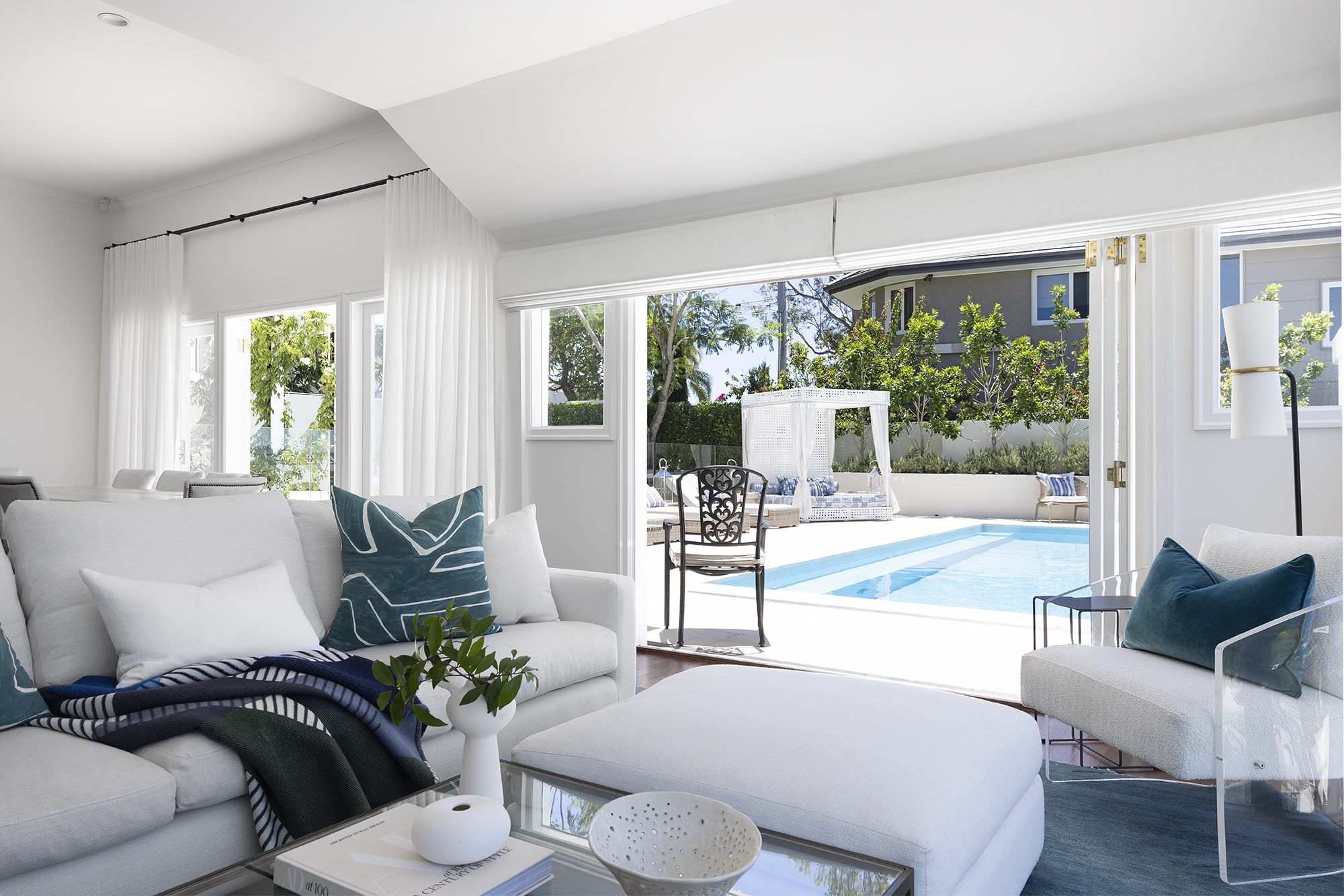 Bright living room interior with plush white sofas, blue-toned cushions and throws, transparent coffee table, and open sliding doors revealing a backyard pool, lounge area, and lush greenery.