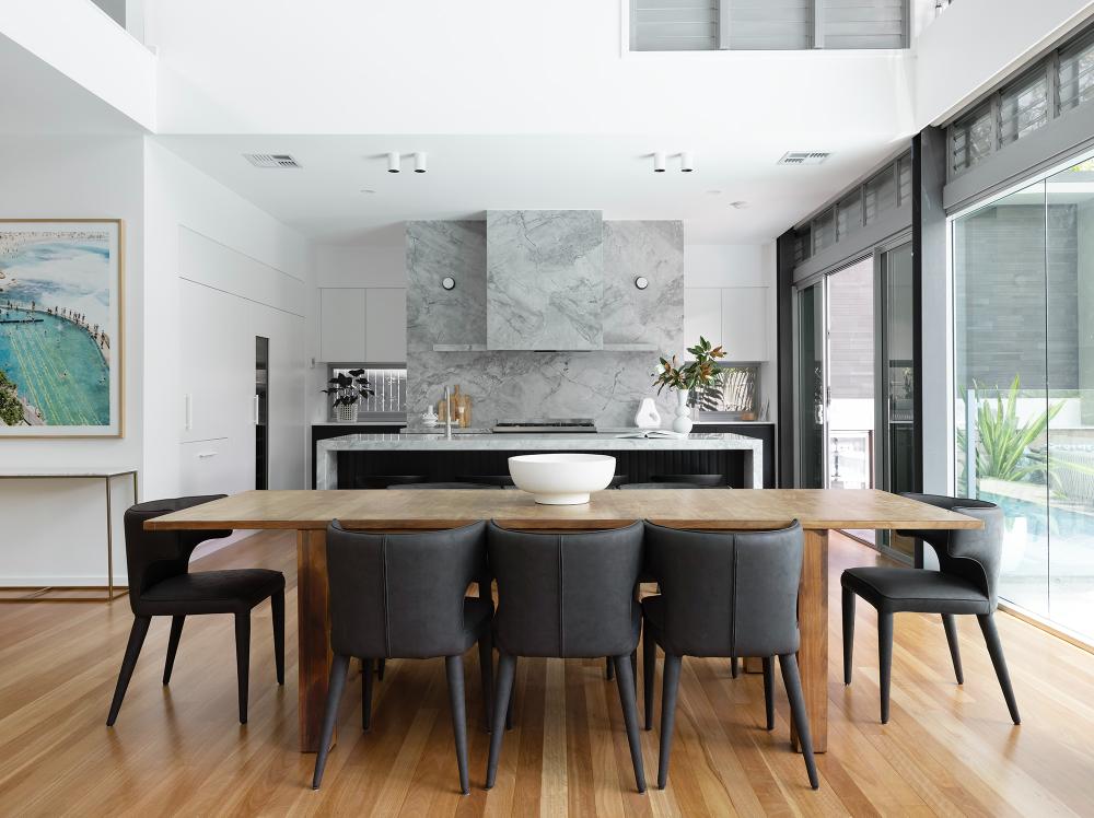Contemporary kitchen with natural wood dining table and black chairs, marble wall feature, and abundant natural light.