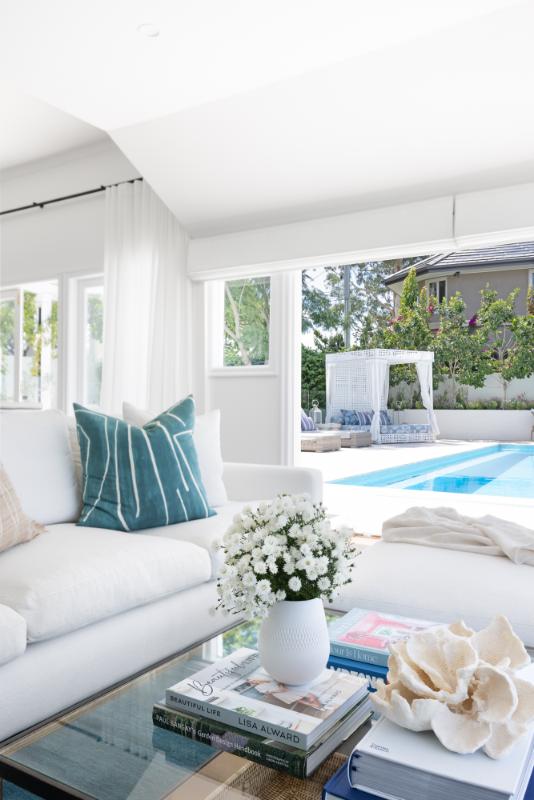 An elegant living room opens up to a serene poolside, featuring white decor with teal and neutral accents, a glass coffee table with decorative books and flowers, and a glimpse of a relaxing outdoor cabana area.