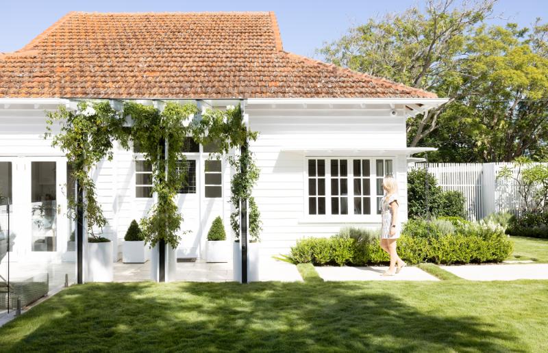 Woman in white standing outside a traditional Queenslander home with greenery and a terracotta roof, embodying Brisbane's charming residential architecture.