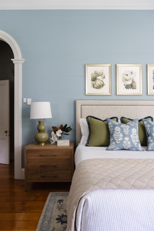 Elegant master bedroom design featuring a soft blue wall, layered bedding in neutrals and greens, and botanical artwork above an upholstered bed — designed by Bella Vie Interiors.